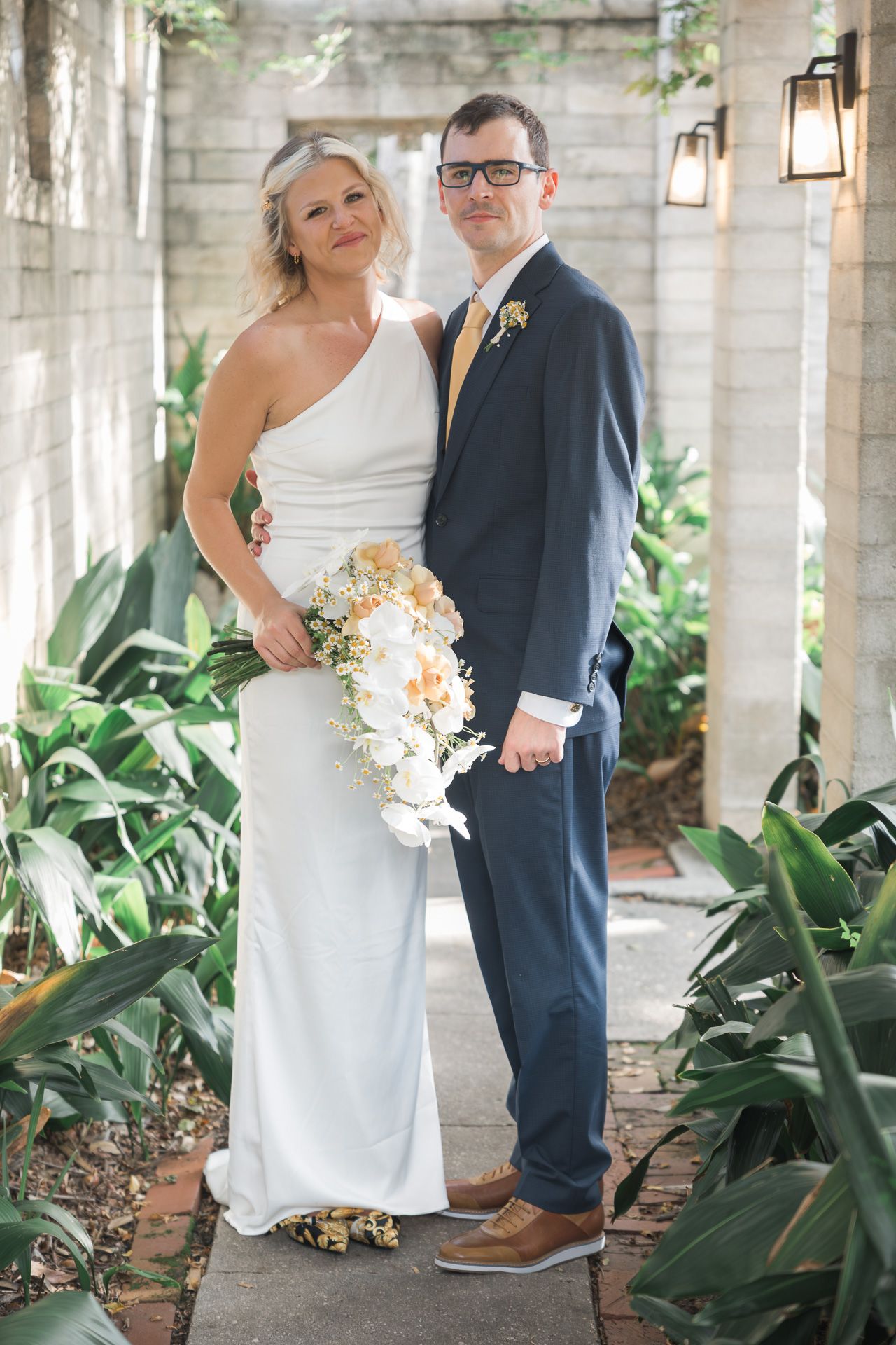 Wedding couple posing in a lush garden walkway with tropical plants and stone columns.
