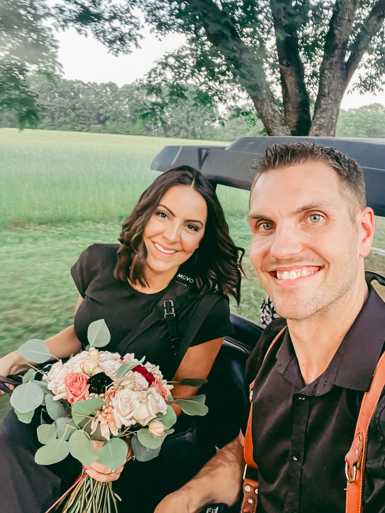 Nick Snell and Brittany Snell sitting on the back of a golf cart smiling holding wedding flowers and camera equipment