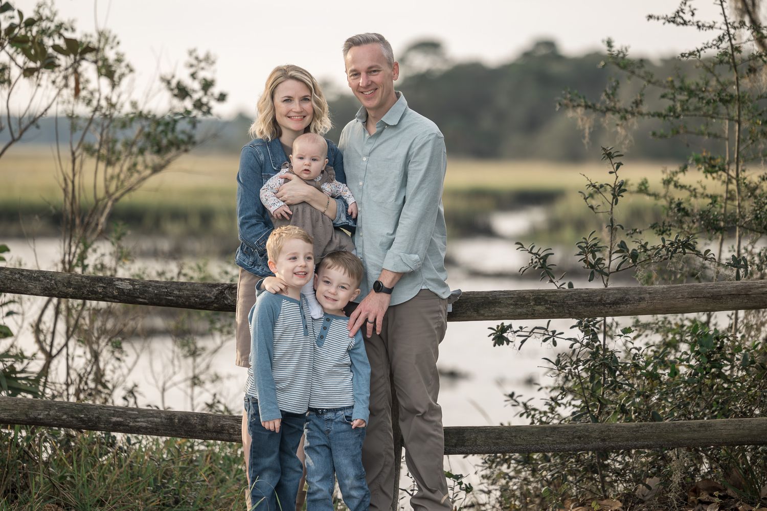 Family portrait session by a wooden fence overlooking a peaceful lake at dusk.