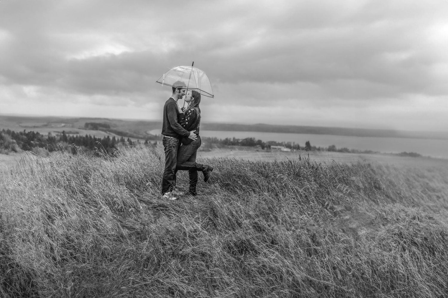 A couple shares an umbrella while standing in a windswept field under moody skies in black and white.