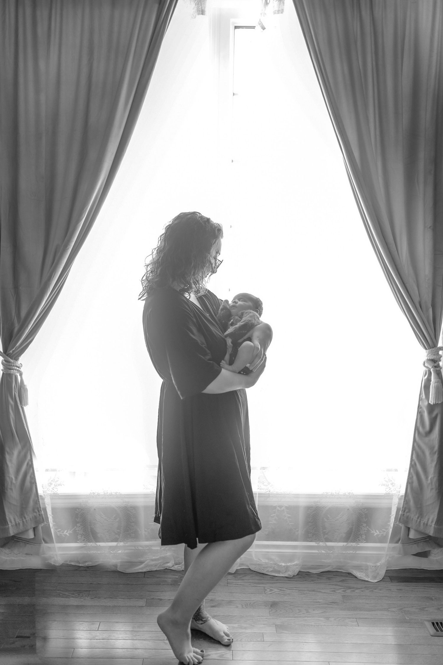A romantic silhouette of motherhood near tall curtained windows in black and white.