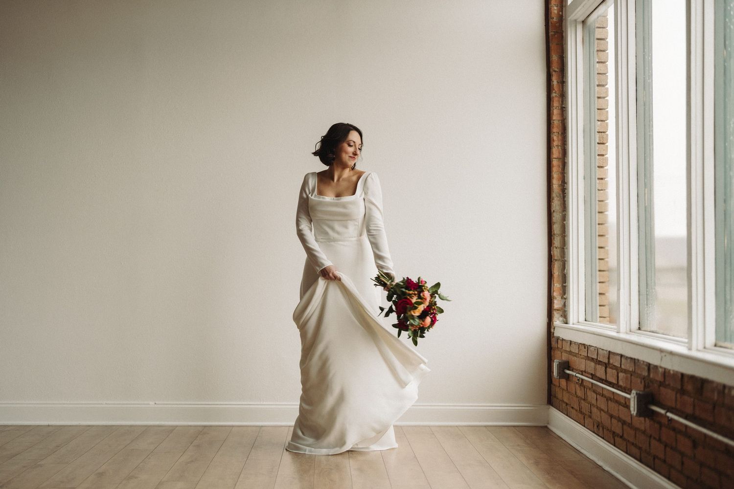 Someone in a flowing white wedding dress stands by a window holding a vibrant bouquet of flowers.