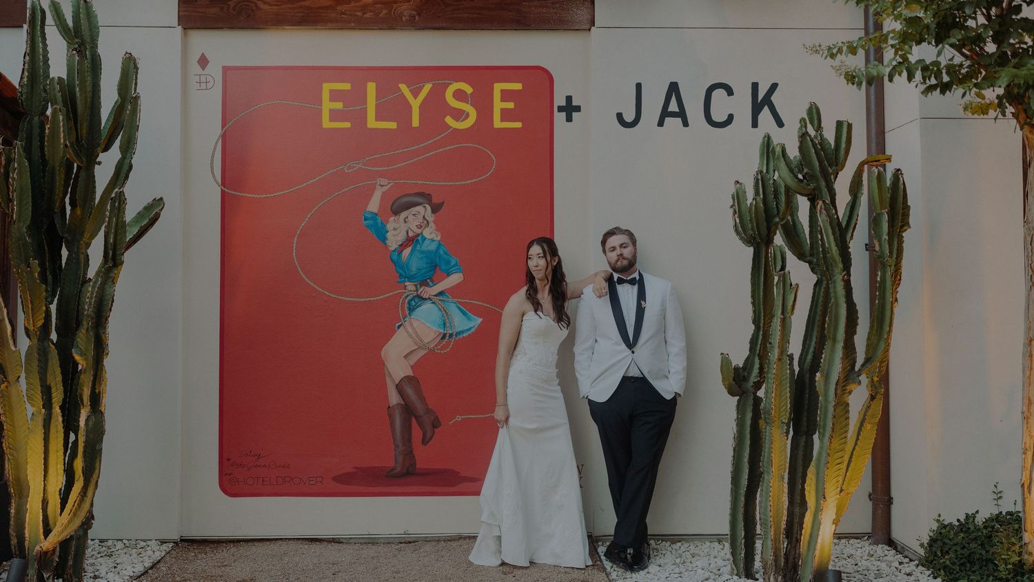 Bride and groom pose in front of a colorful Elyse and Jack wedding sign with cacti outdoors.