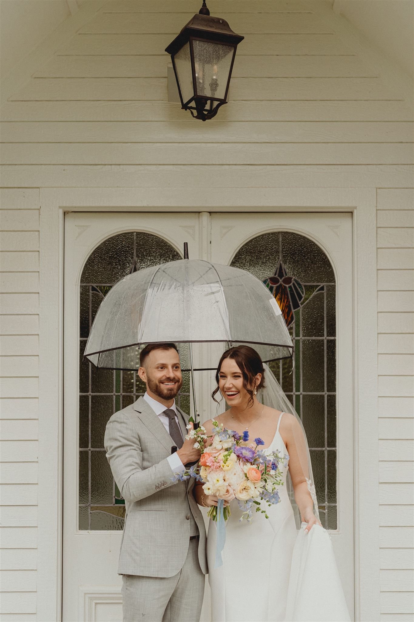 A romantic wedding couple shelters under a clear umbrella in front of a white chapel door with a vintage lantern above.