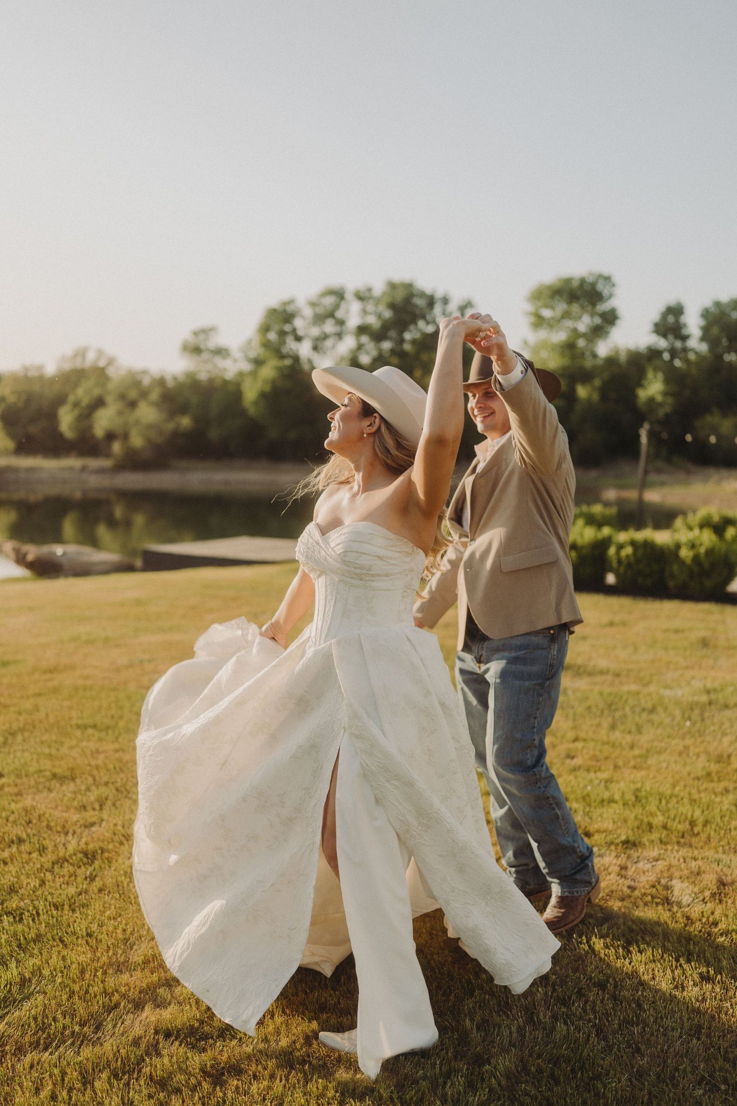 A couple dancing together on a grassy field at sunset with a lake in the background during a wedding celebration.