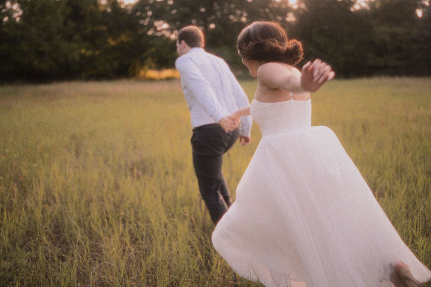 Two people in wedding attire stand close together in a sunlit golden field during sunset.
