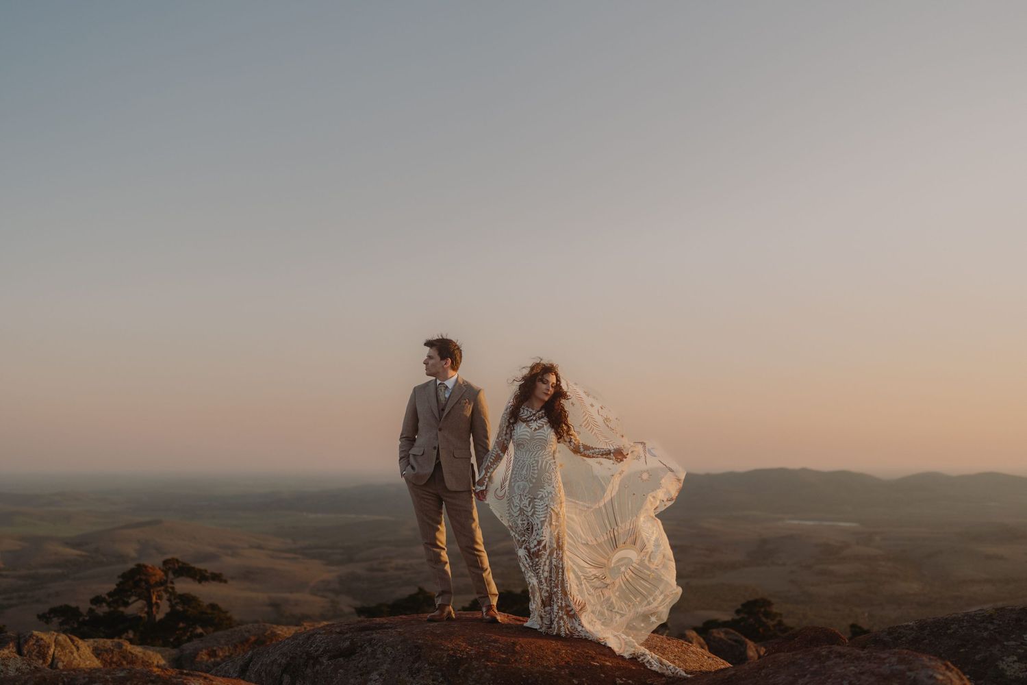 Couple stands on mountain peak at sunset while bride's flowing white dress catches the wind in dramatic pose.