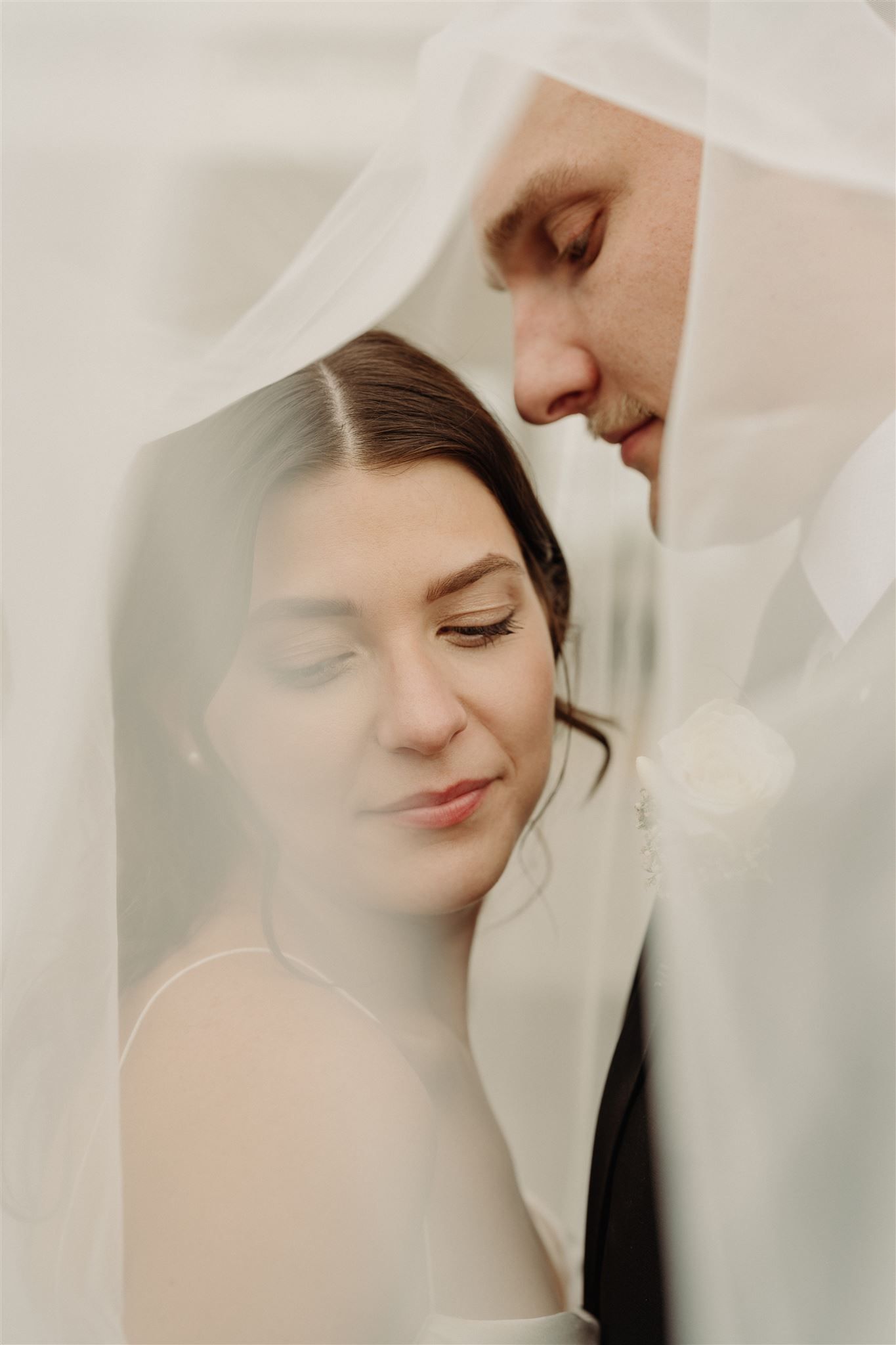 Intimate wedding photography of couple beneath soft white veil in romantic ethereal lighting.