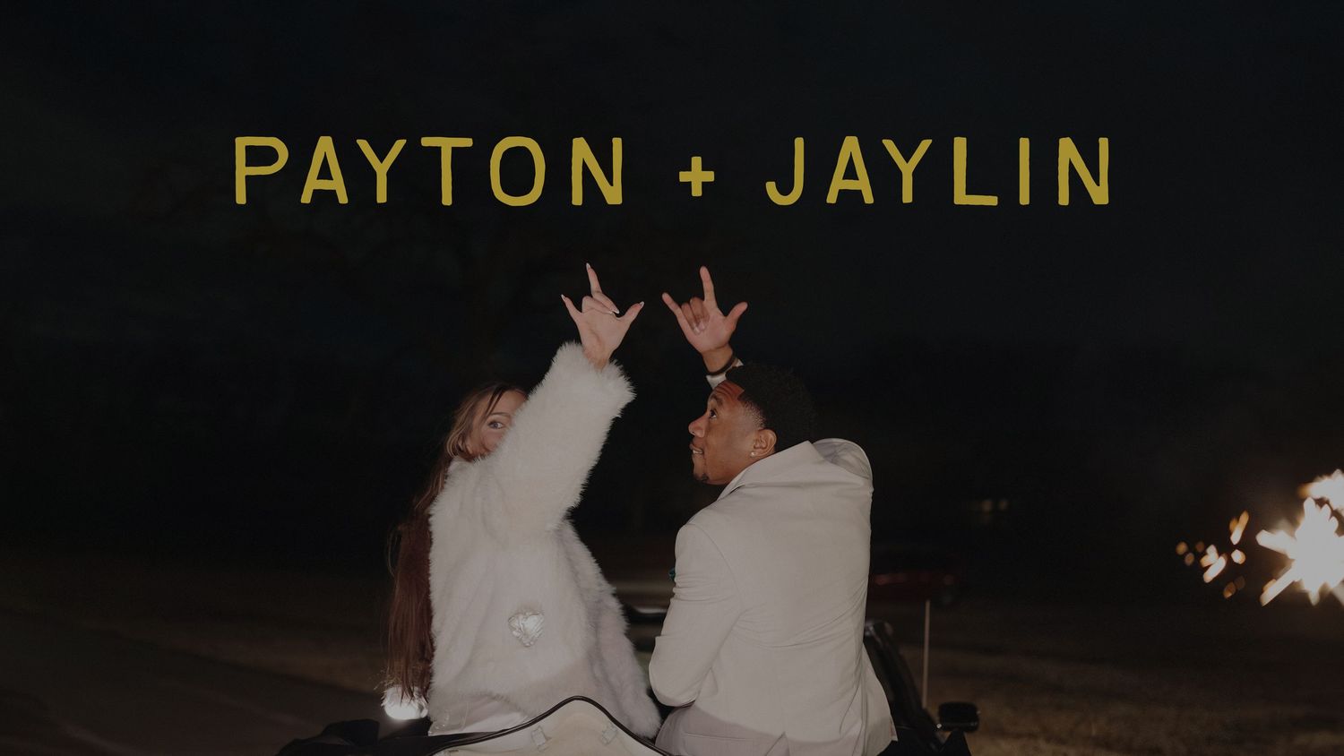 Payton and Jaylin couple sitting by a campfire at night, making hand signs in matching white hoodies.