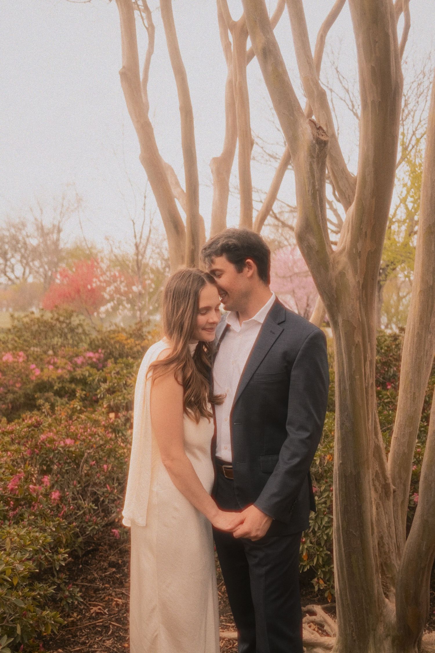 Wedding couple embraces in front of autumn trees and pink flowers during outdoor photo session.