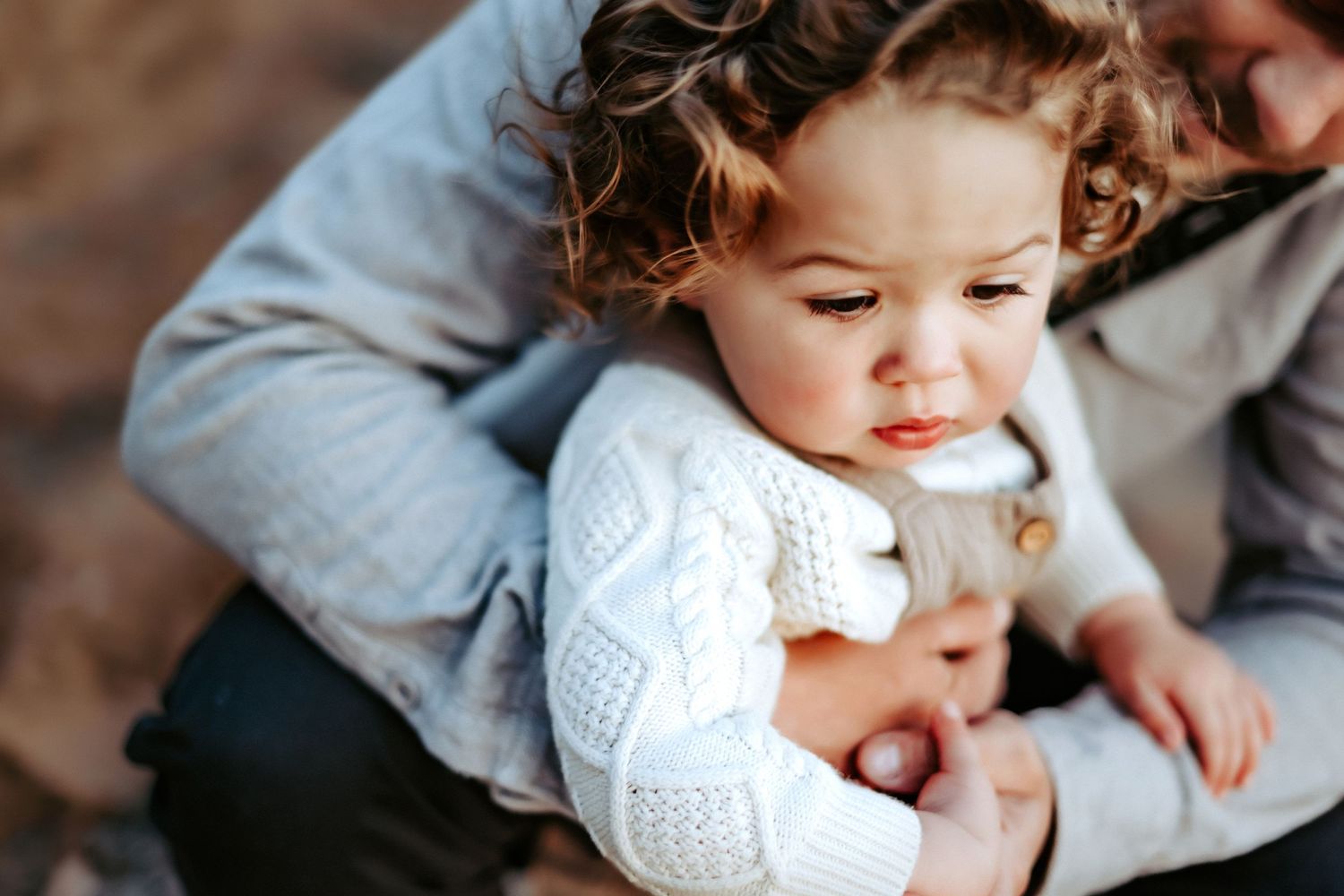 A small child in a white knit sweater is held in someone's lap.