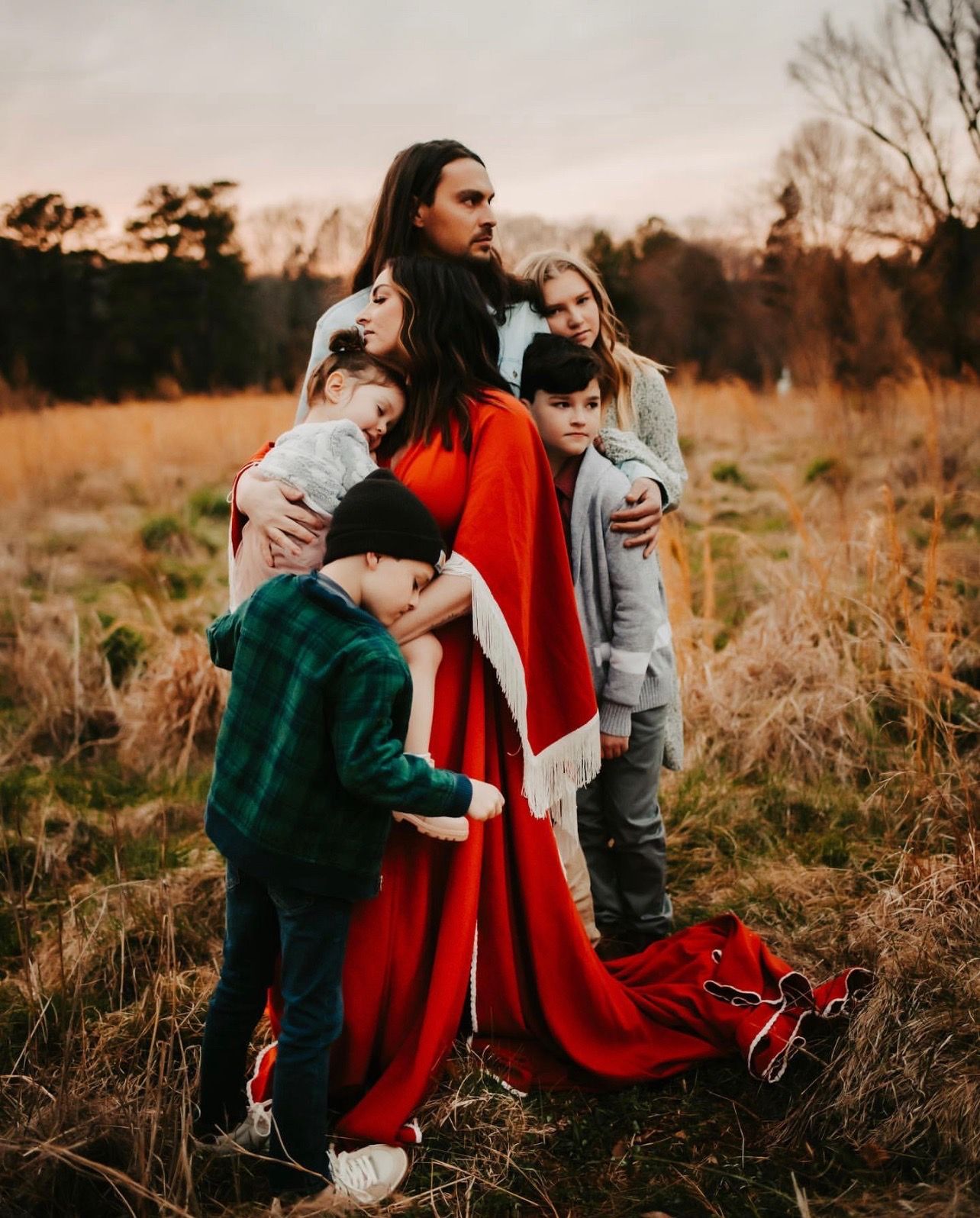 Family wearing winter clothing embraces together in golden field at sunset.