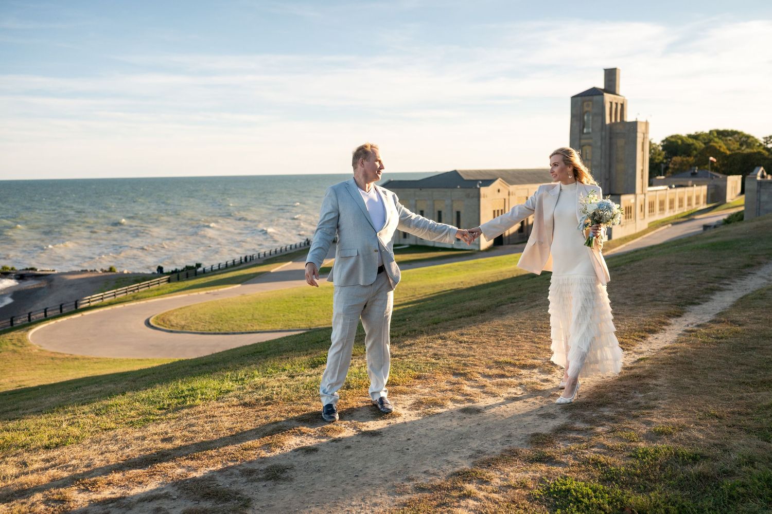 Bride and groom hold hands walking on a grassy coastal path with ocean and historic building backdrop.
