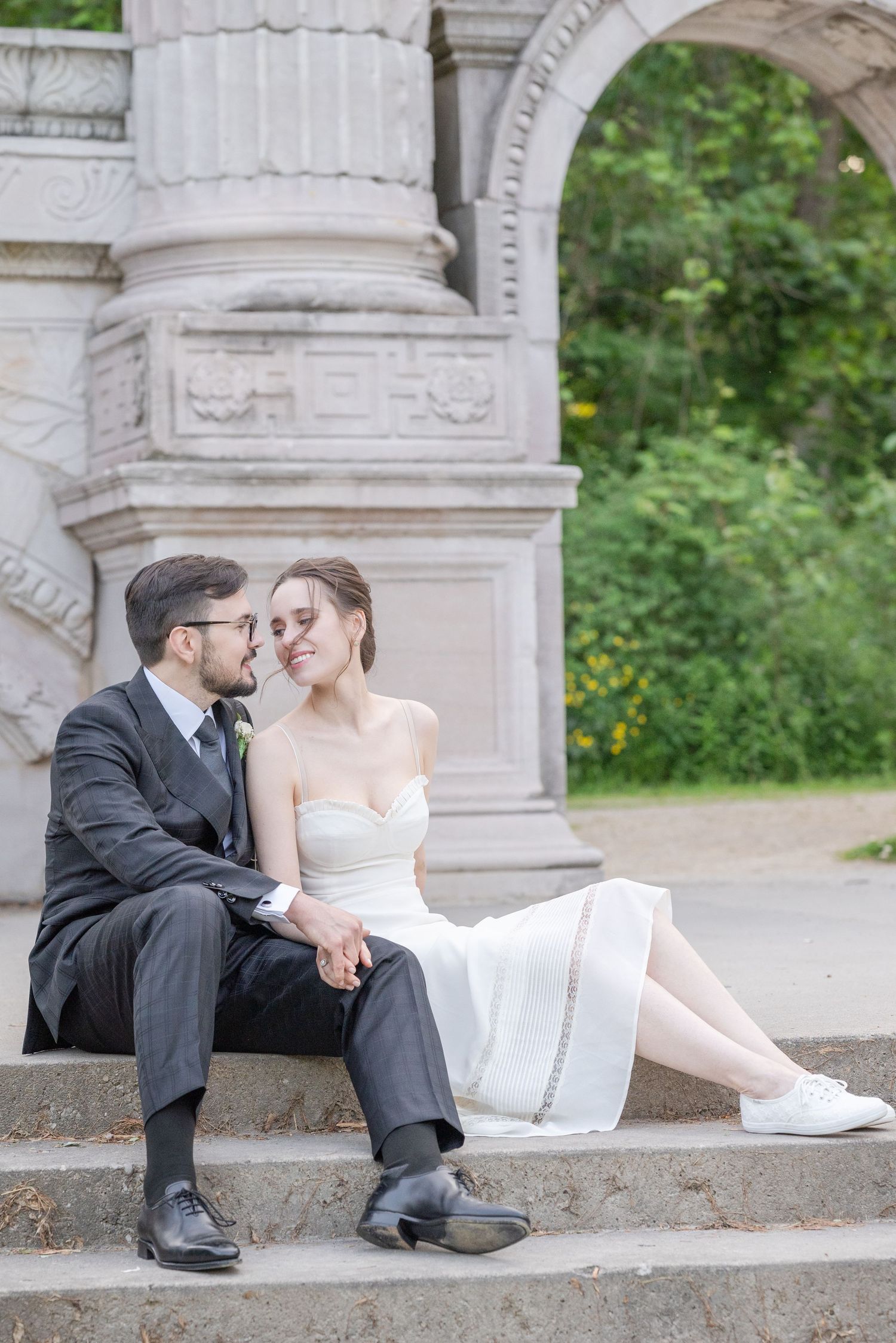 Bride and groom sit on stone steps under ornate arch, sharing a tender moment in white dress and gray suit.