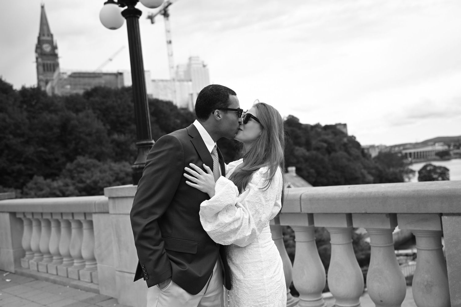 Black and white photo of an interracial couple sharing a romantic kiss on a bridge with a historic cityscape behind them.