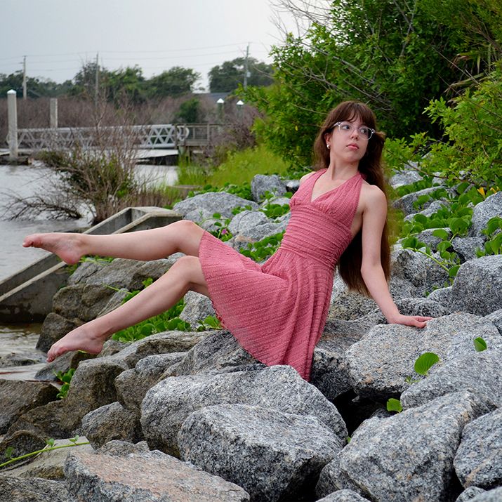 Young woman in a coral pink dress and glasses sits barefoot on large gray rocks beside the water on a cloudy day.
