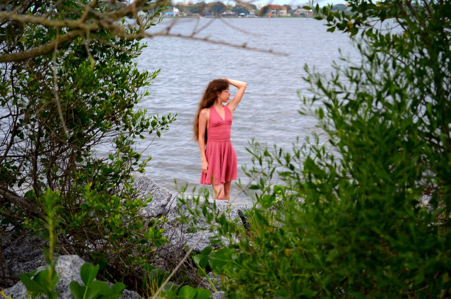 Woman in a pink dress poses among green foliage at the water’s edge with one hand resting on her head.