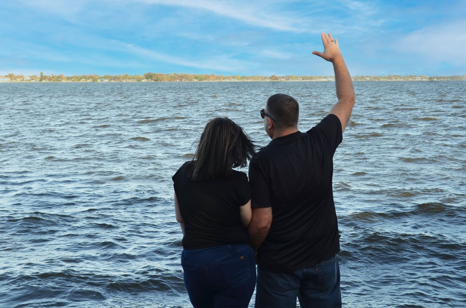 Couple stands with their backs to the camera beside a lake as the man waves toward the distant shore.