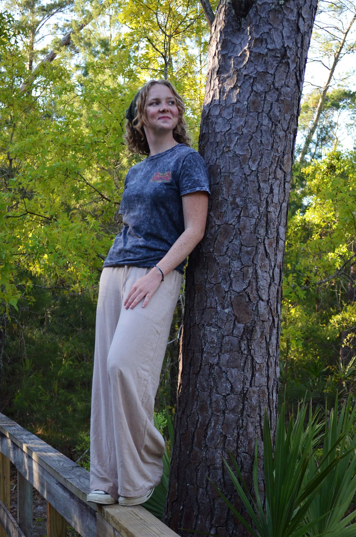 oung woman in a gray T-shirt and tan pants leans against a pine tree on a wooden walkway surrounded by green foliage.