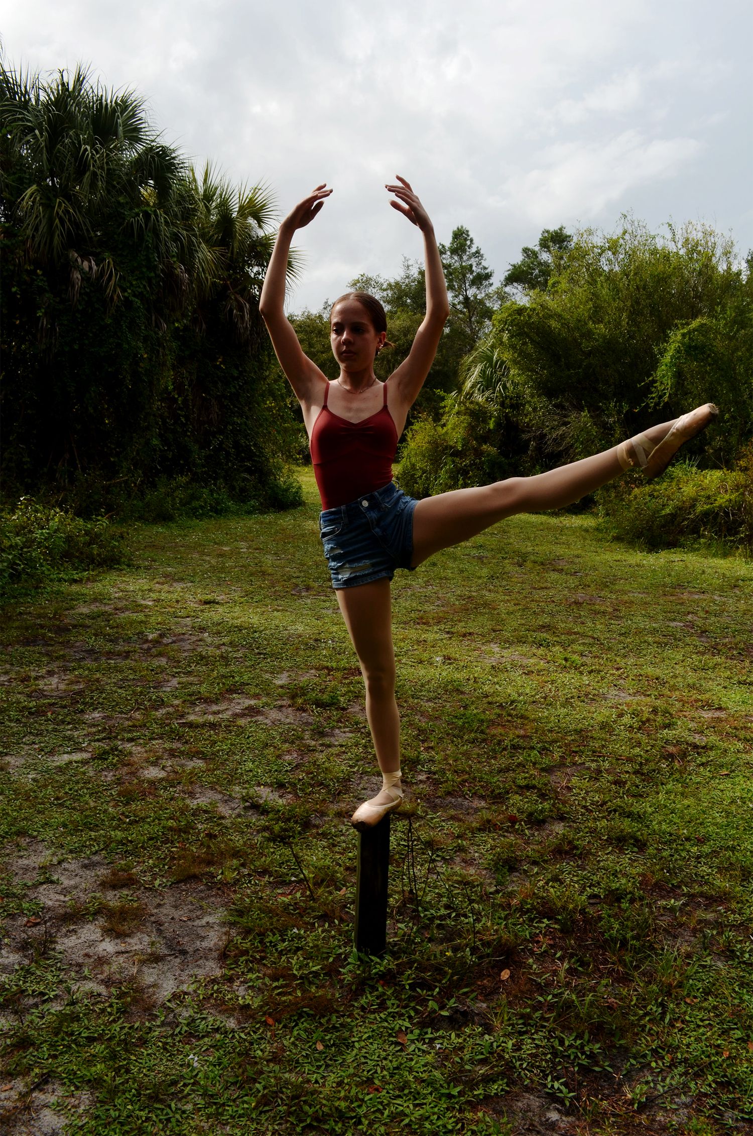 Ballet dancer in denim shorts and pointe shoes balancing on a post with one leg extended high in a grassy field.