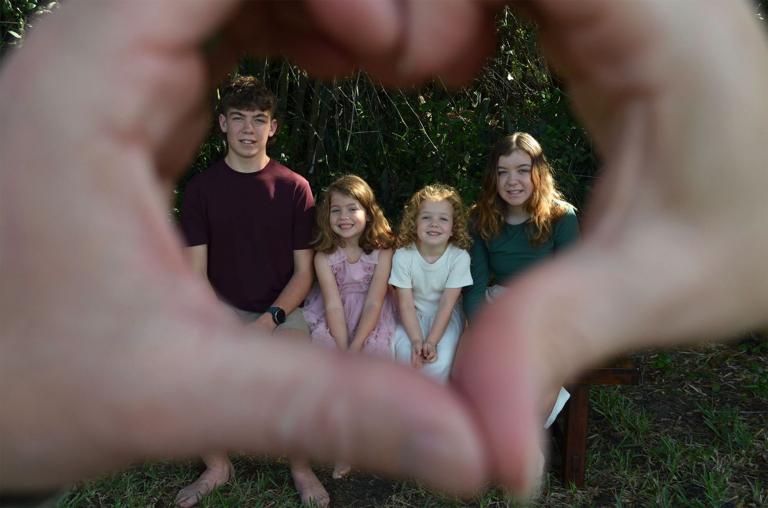 Four children, a teenage boy and three girls, smiling outdoors through a heart shape formed by hands in the foreground.
