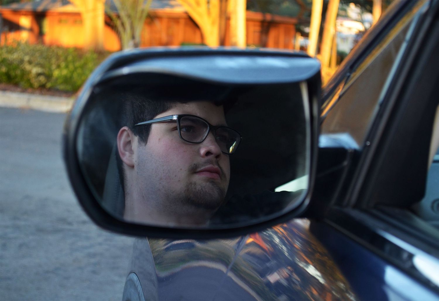 Man wearing glasses reflected in a car’s side mirror while sitting in the driver’s seat on a quiet residential street.