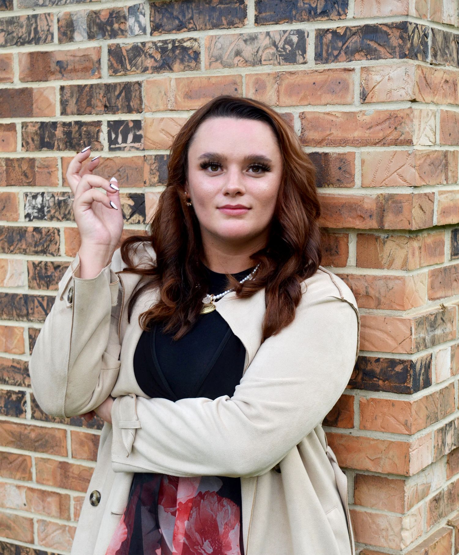 Woman with wavy auburn hair poses in a beige trench coat over a floral outfit, raising one hand near a brick wall.