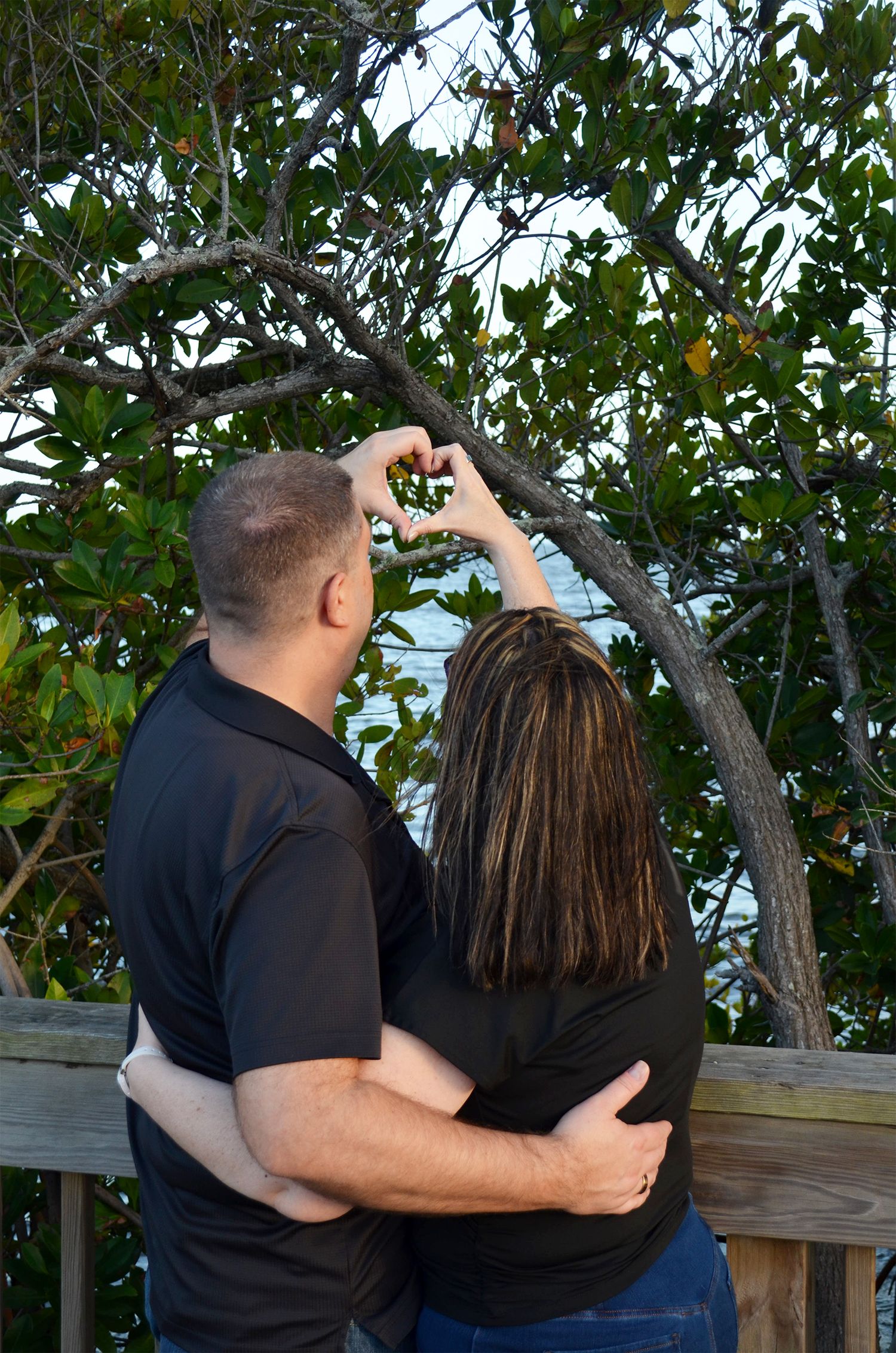 Couple embracing in front of a wooden railing, making a heart shape with their hands beneath a leafy tree.