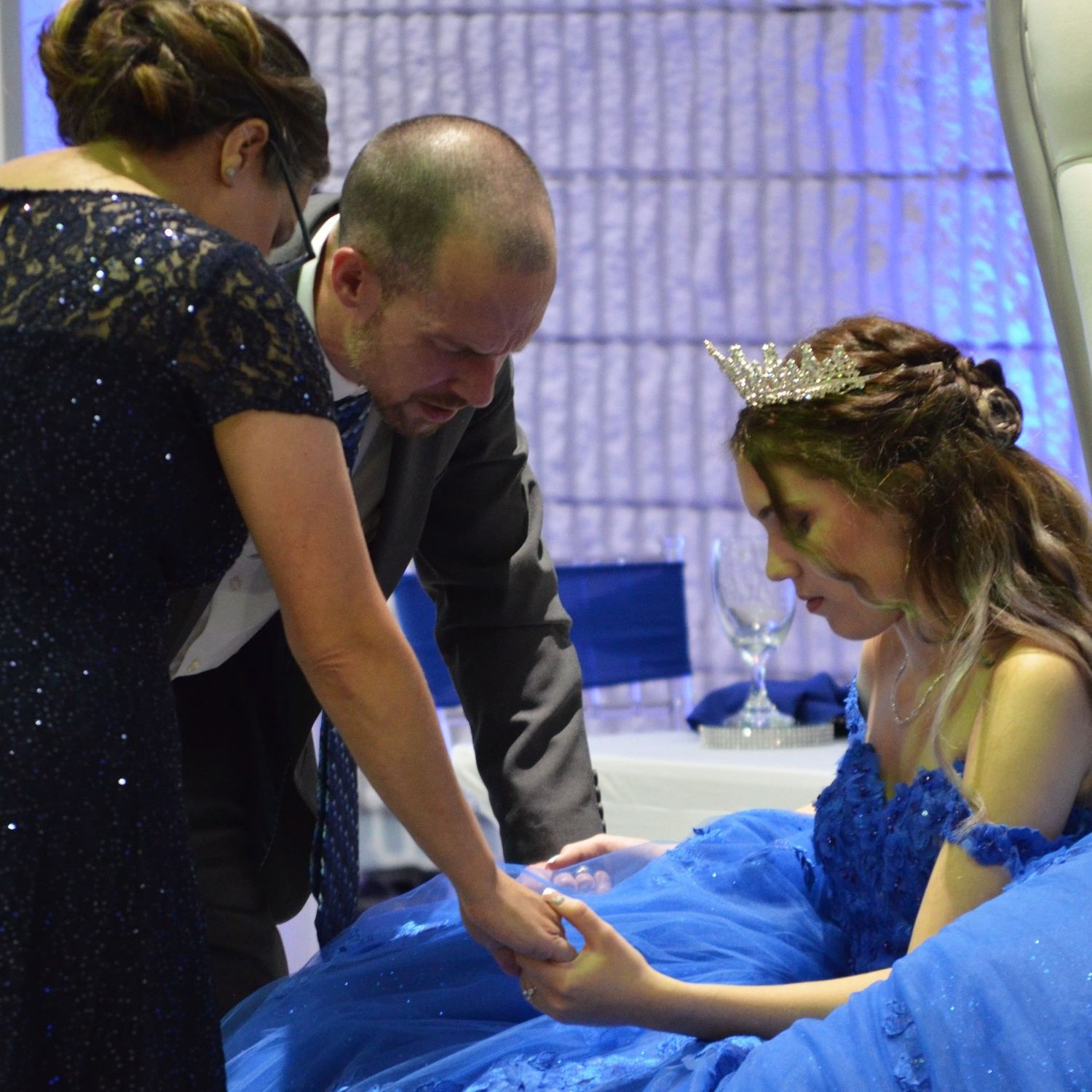 Girl in a blue gown and tiara seated at a celebration as a man and woman in a sparkling dress lean in to hold her hand.