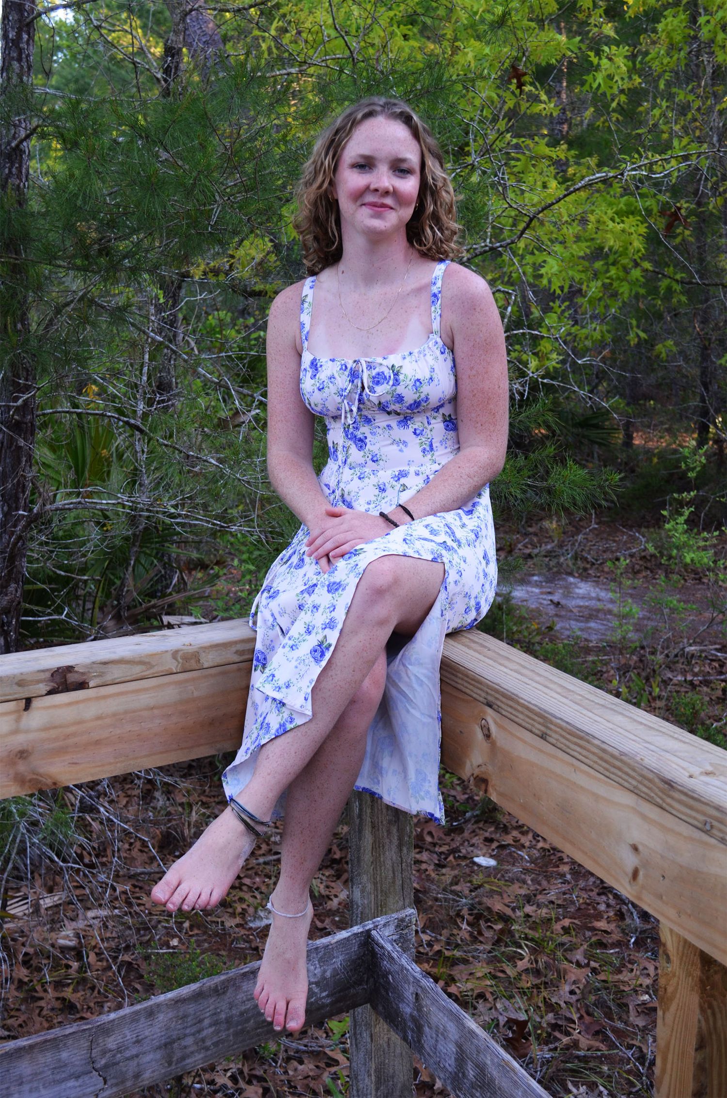 Young woman in a blue floral sundress and ankle bracelet sits barefoot on a wooden railing in a sunlit pine forest.