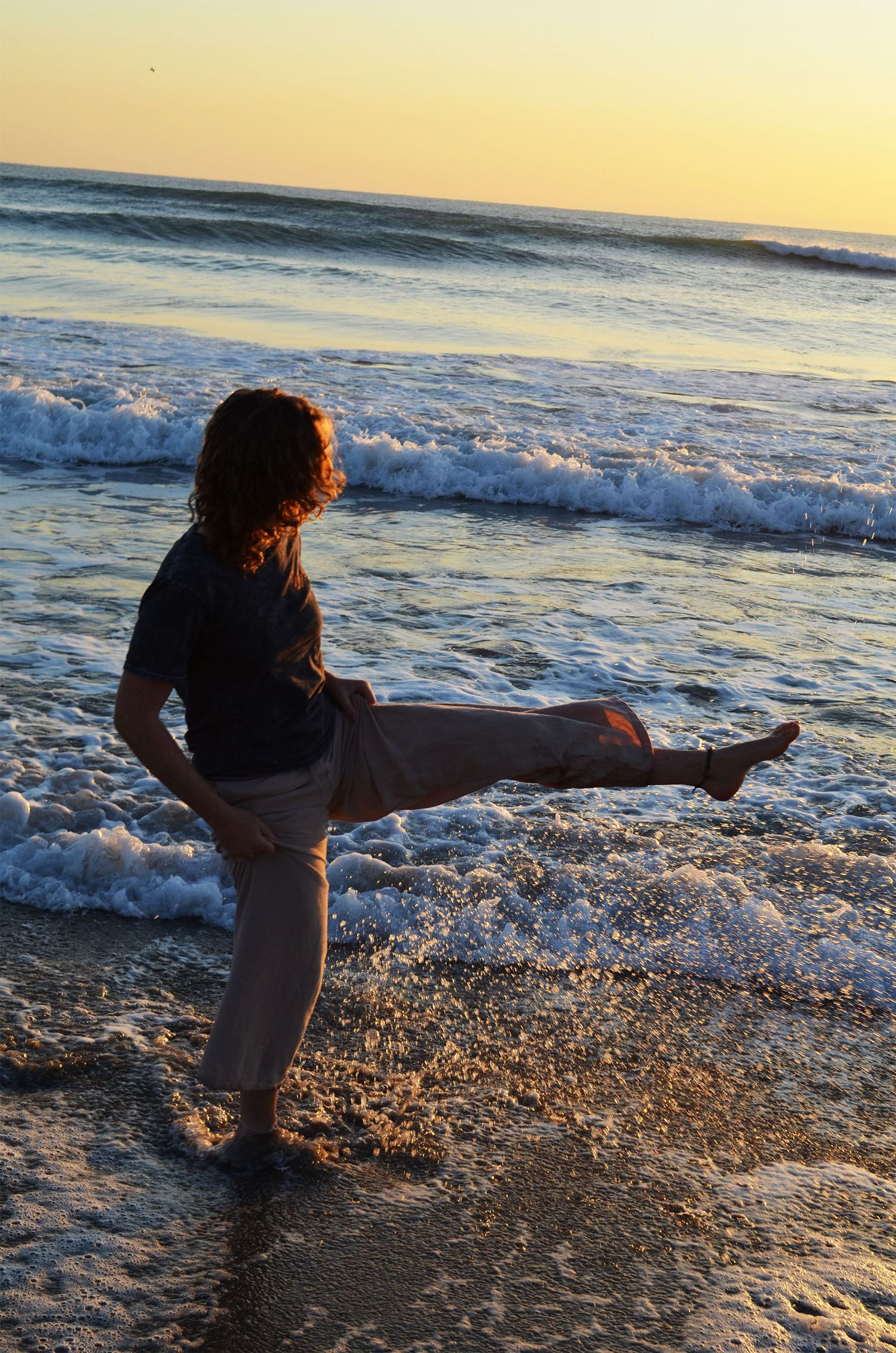 Person in a dark shirt and loose pants kicks one leg high on wet sand as waves roll in at sunrise.