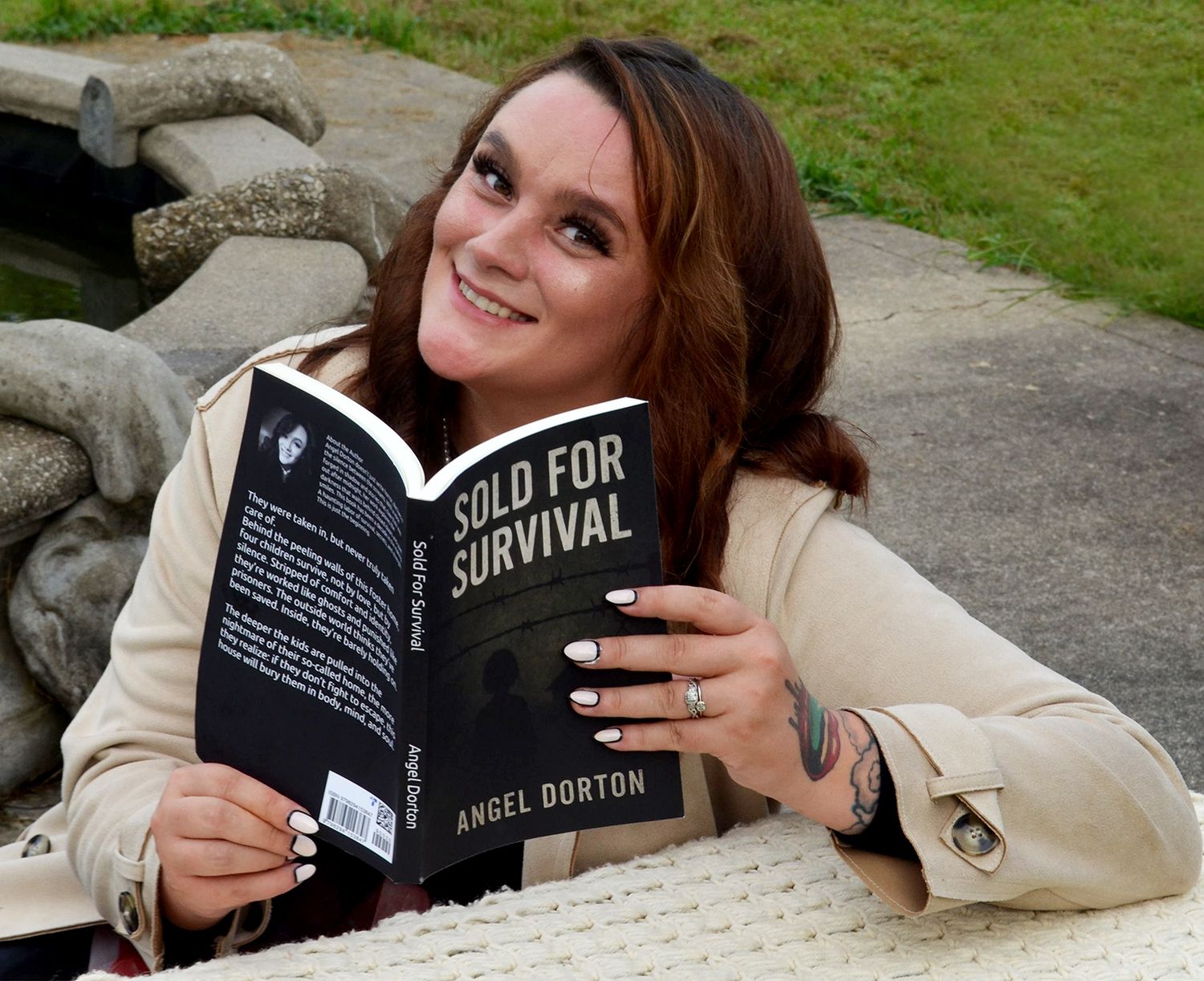 Smiling woman with long auburn hair holds Sold for Survival by Angel Dorton while seated at an outdoor table.