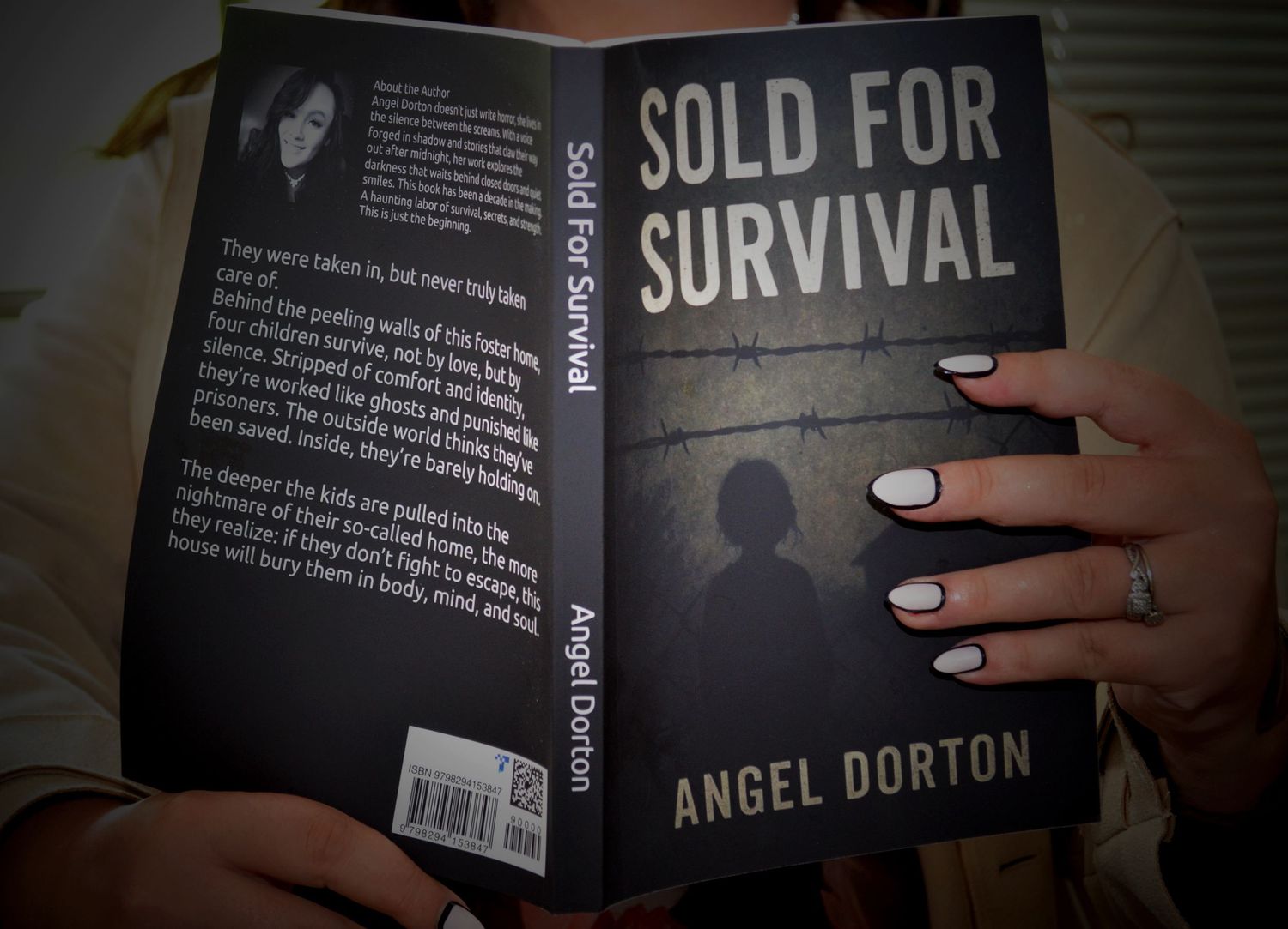 Woman with white-tipped manicured nails holds up the dark cover of Sold for Survival by Angel Dorton.