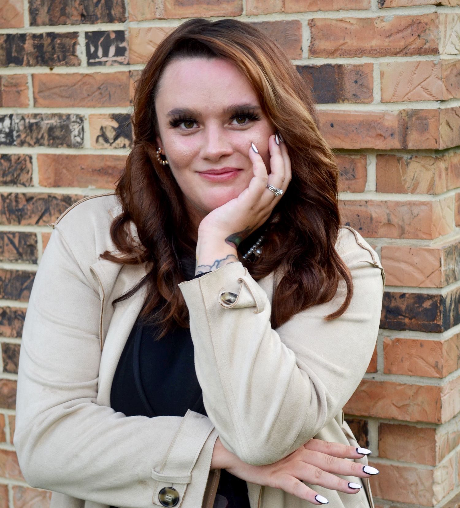 Smiling woman with long auburn hair rests her chin on her hand while posing in a beige trench coat against a brick wall.