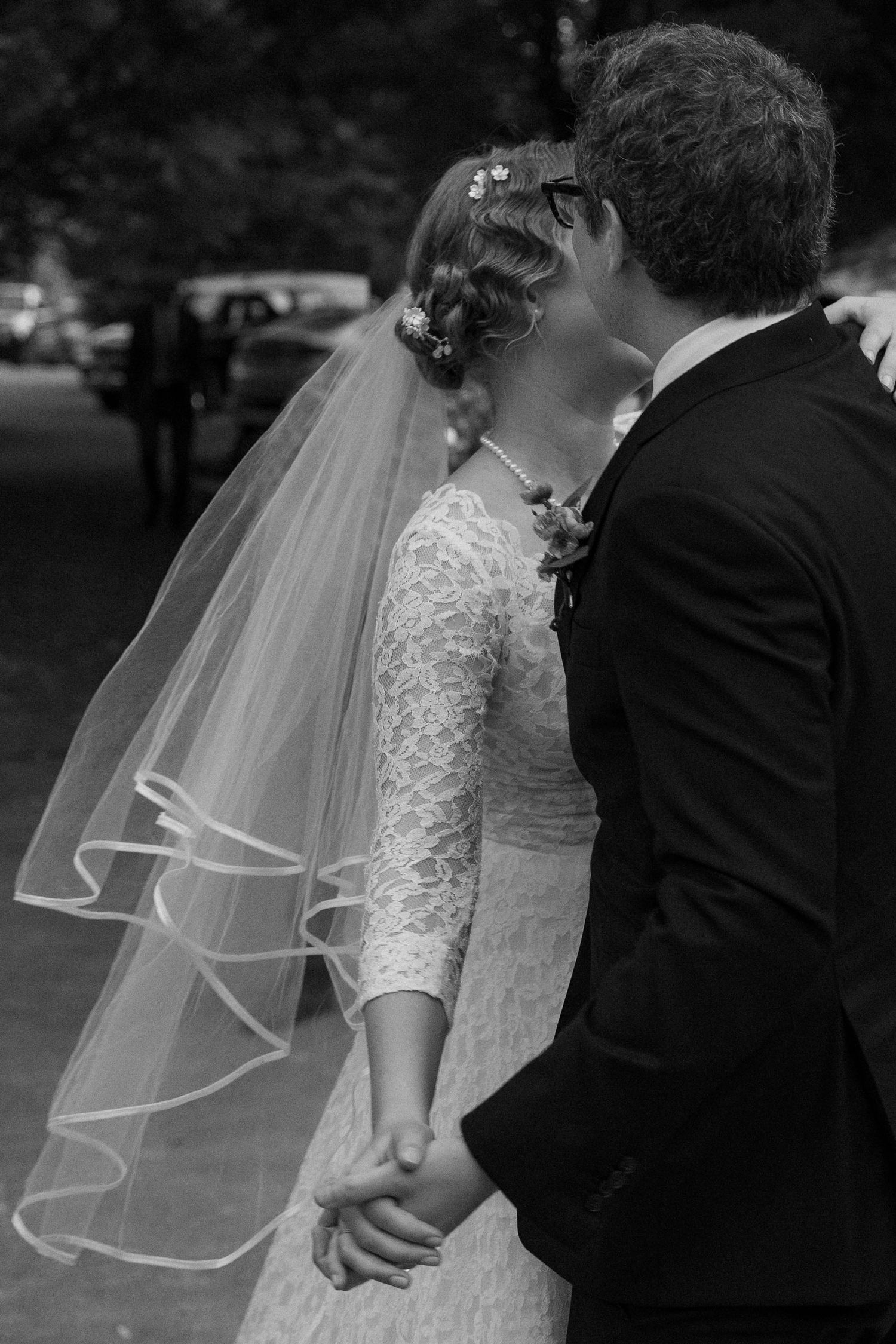 Black and white photo of a bride in a lace gown and veil kissing her groom while holding hands outdoors.