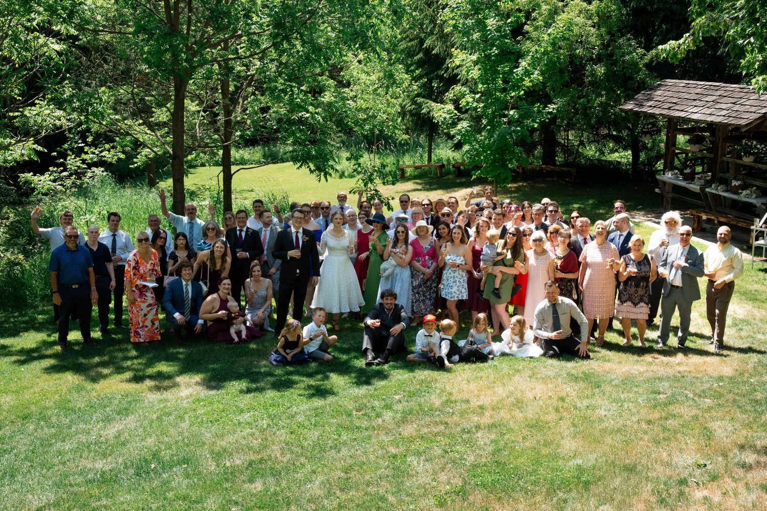 A large group of wedding guests poses with the bride and groom on a sunny lawn surrounded by trees and a wooden pavilion.