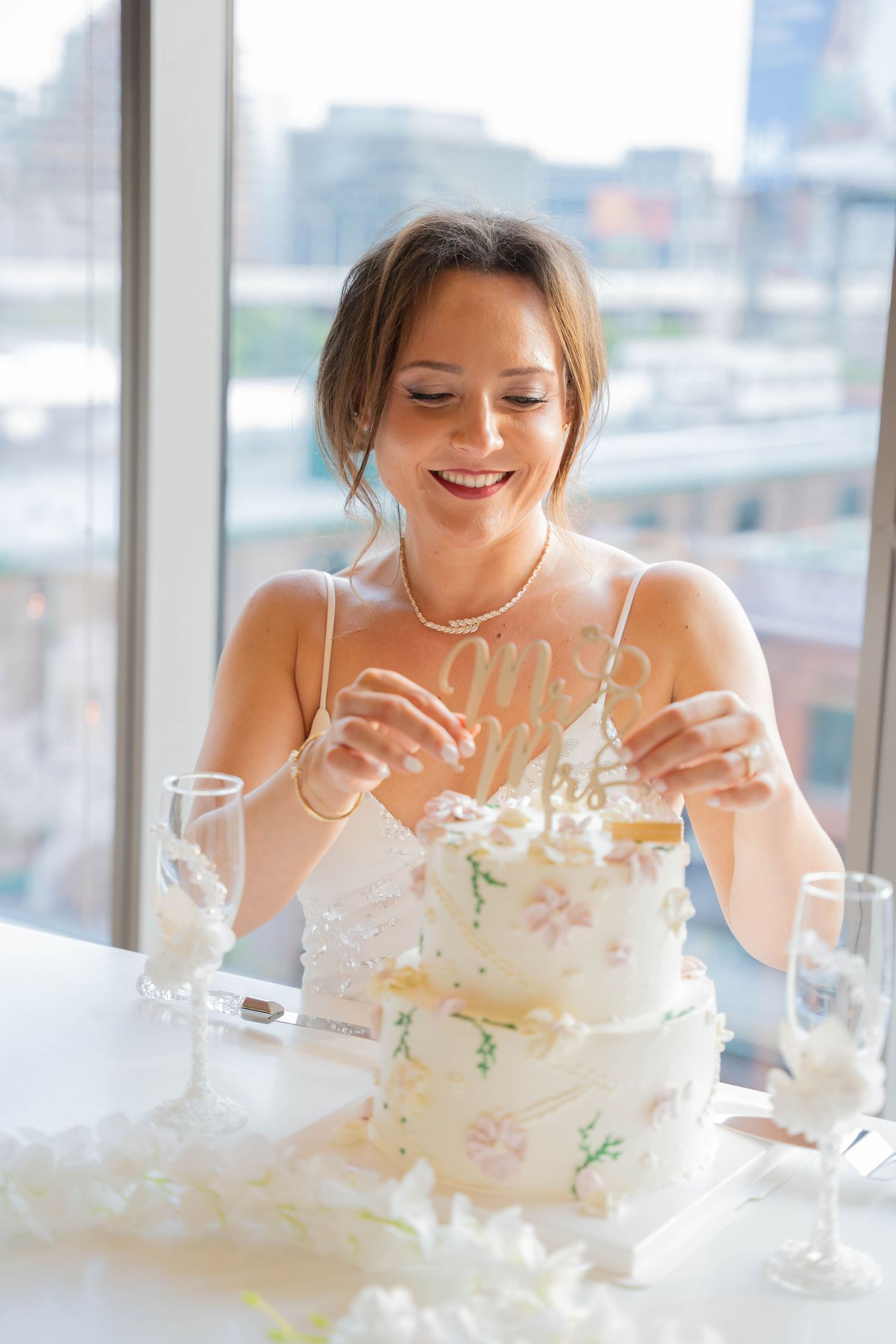 Smiling bride in a white dress adjusts the topper on a two-tier wedding cake beside champagne flutes with a city view behind her.