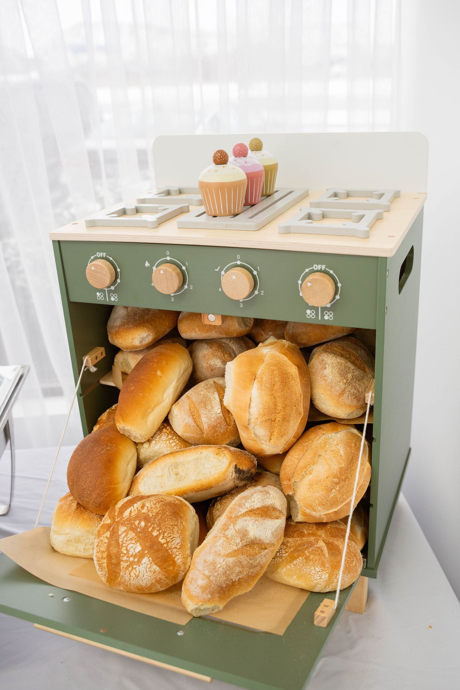 A green toy oven display overflowing with fresh bread rolls, topped with pastel cupcakes at a baby shower.