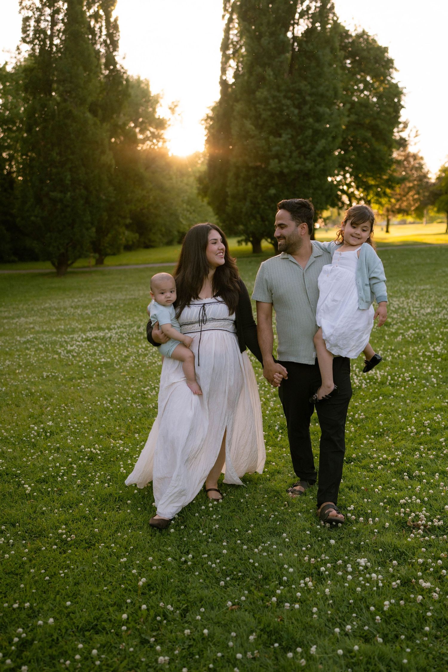 Parents carrying a baby and a young girl stroll across a sunlit grassy field dotted with white clover flowers.