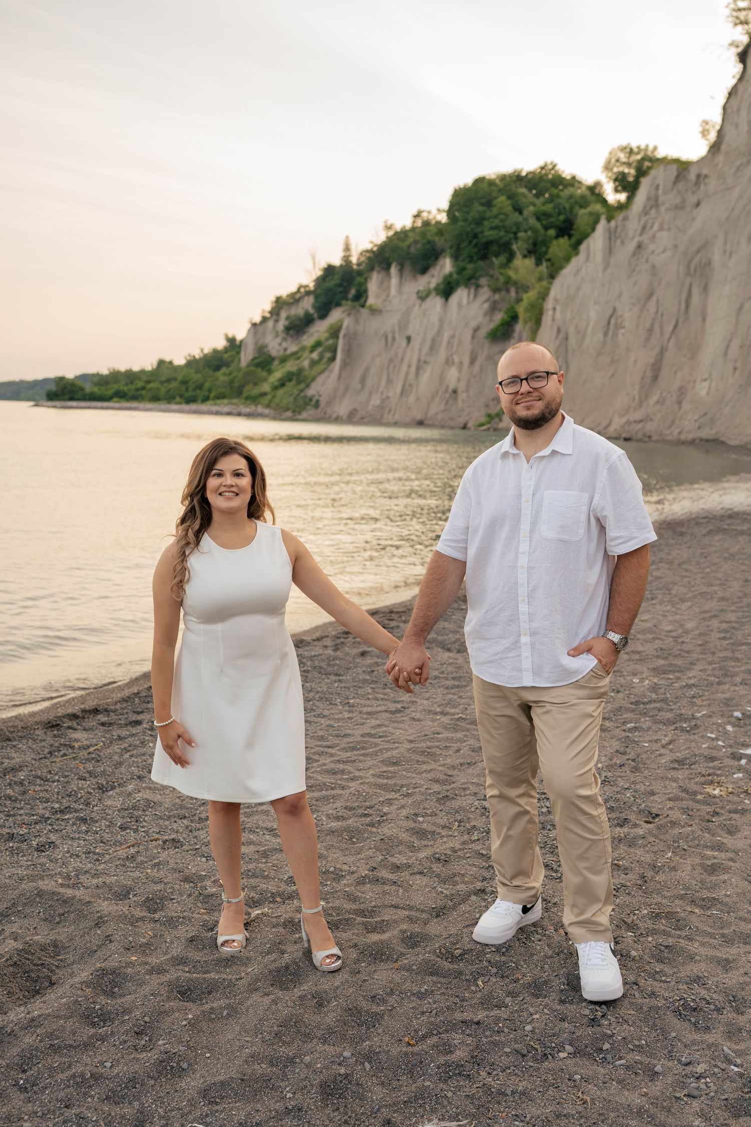 Engaged couple in white outfits holding hands and smiling on a pebble beach with scenic bluffs and calm water behind.