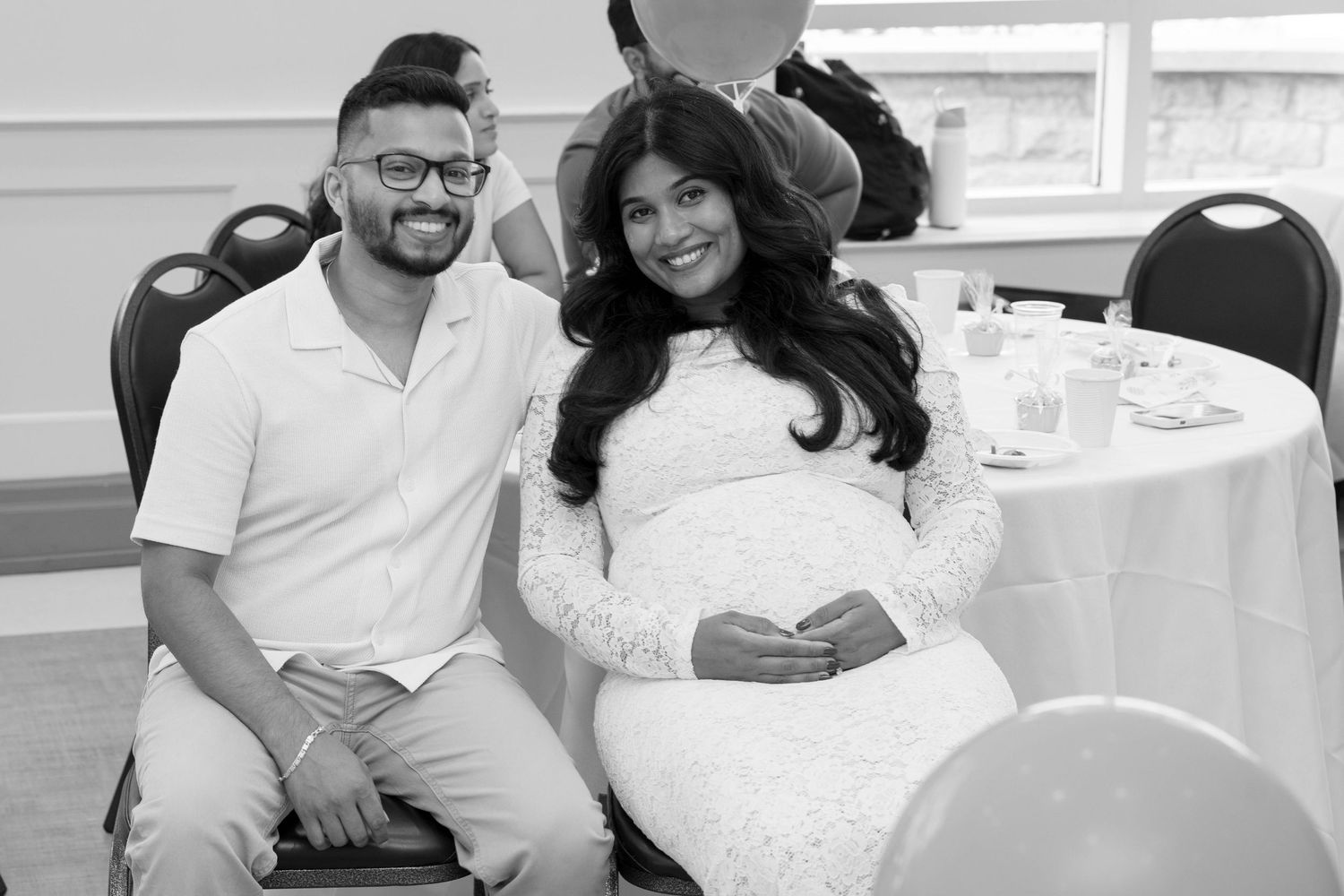 Black and white photo of a smiling expectant couple seated together at a decorated baby shower table.