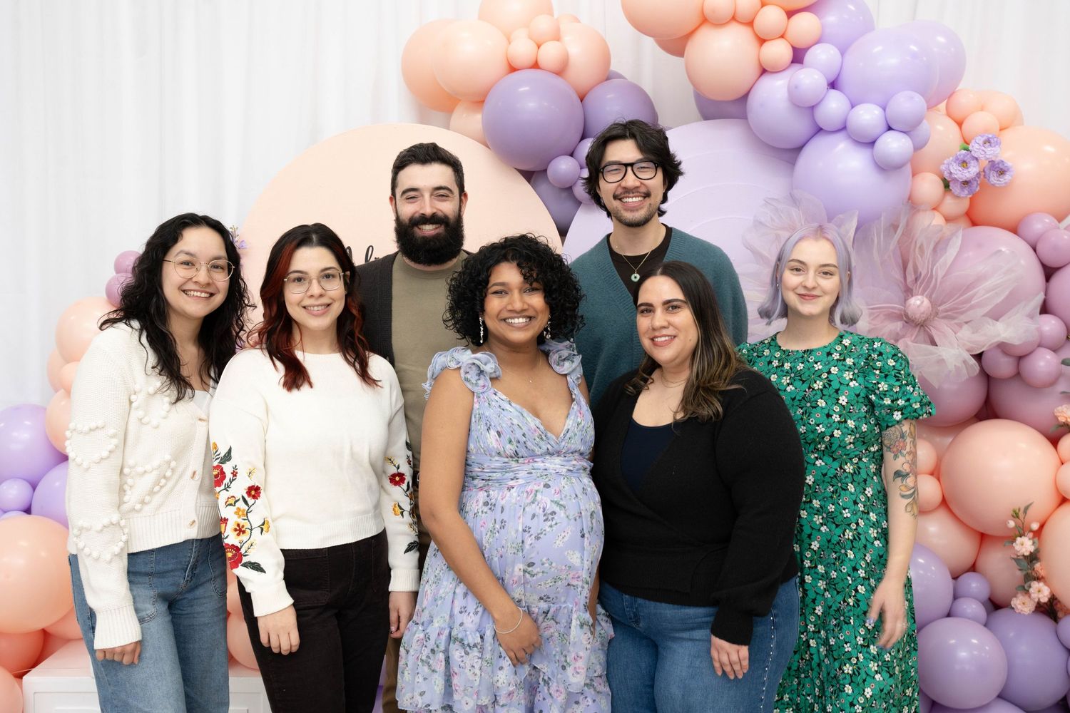A group of seven friends and family smile together beside pink, peach and purple balloons at a baby shower celebration.