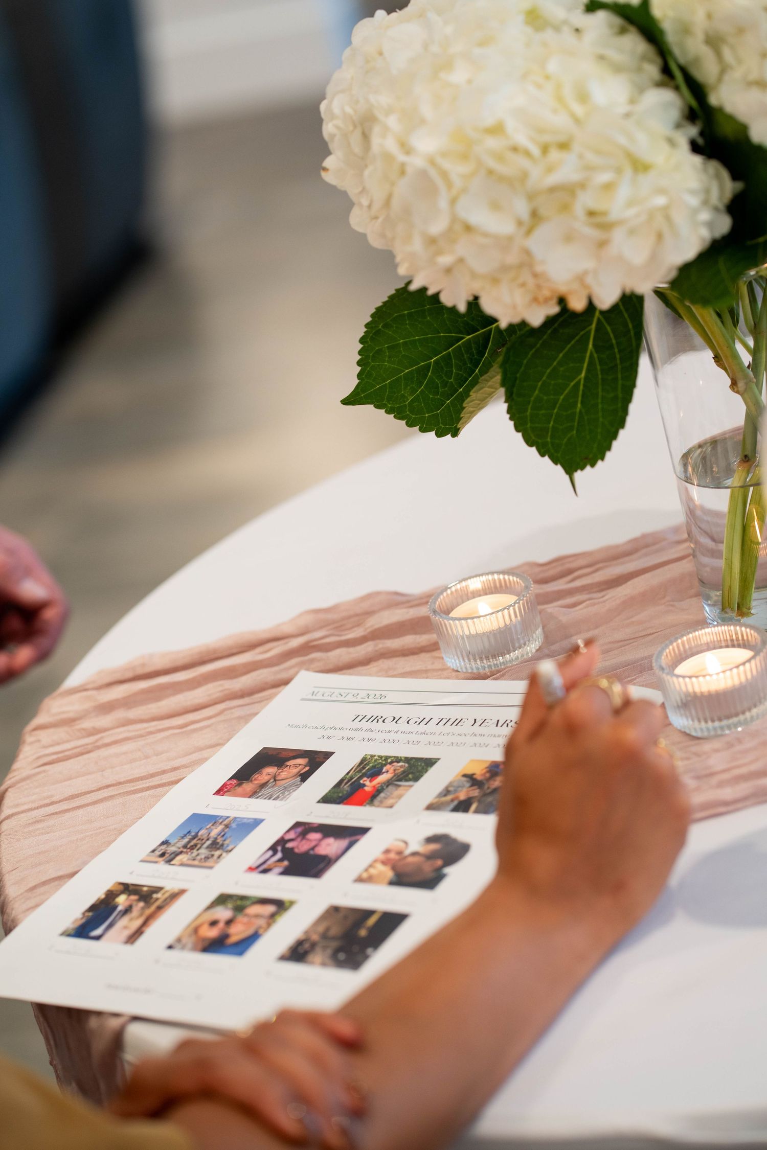 A hand rests on a printed Through the Years photo sheet beside a white hydrangea bouquet and flickering tea lights on a pink table runner.