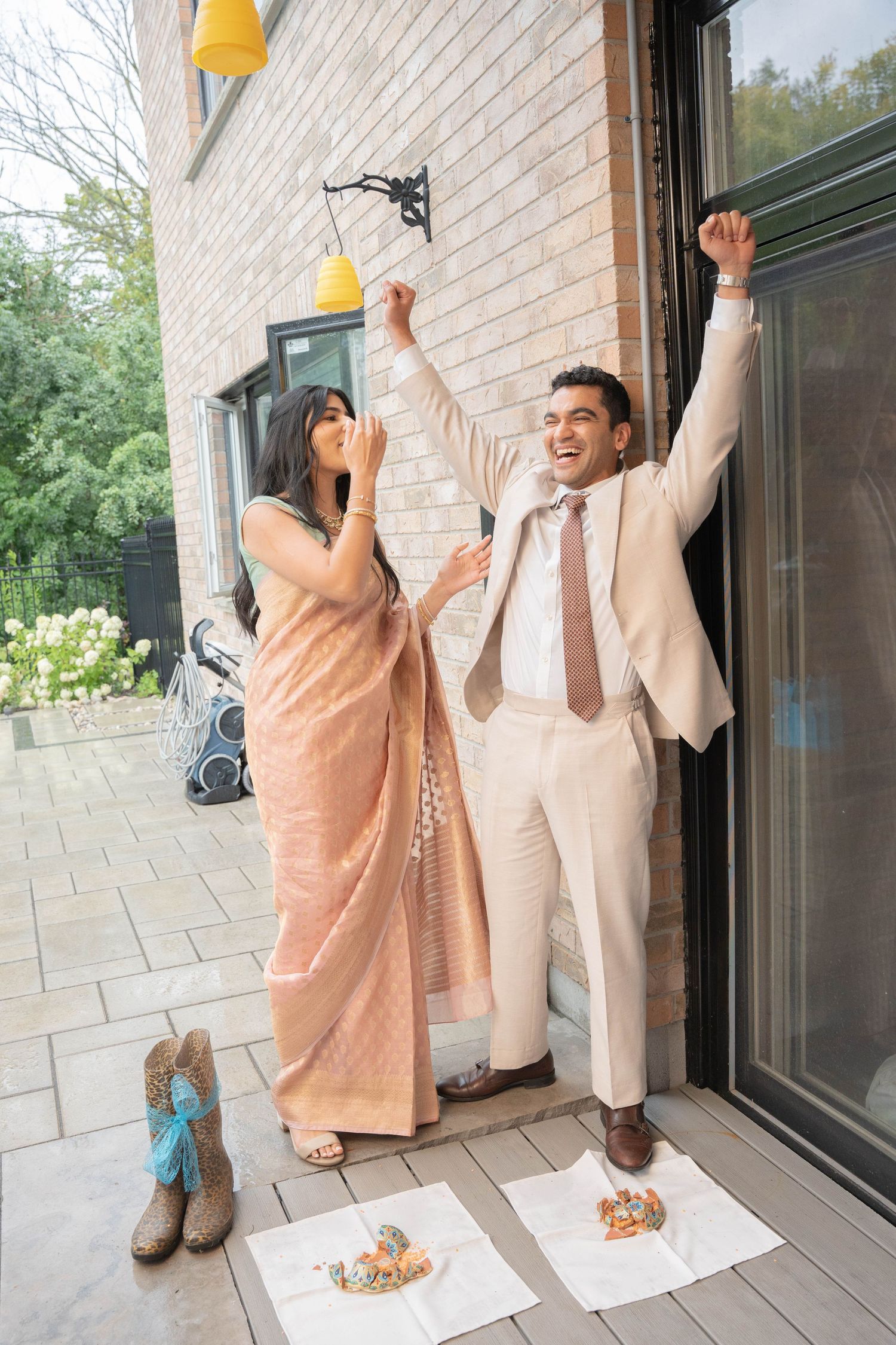 A joyful groom in a beige suit raises his arms in triumph beside his bride in a peach saree on a patio.
