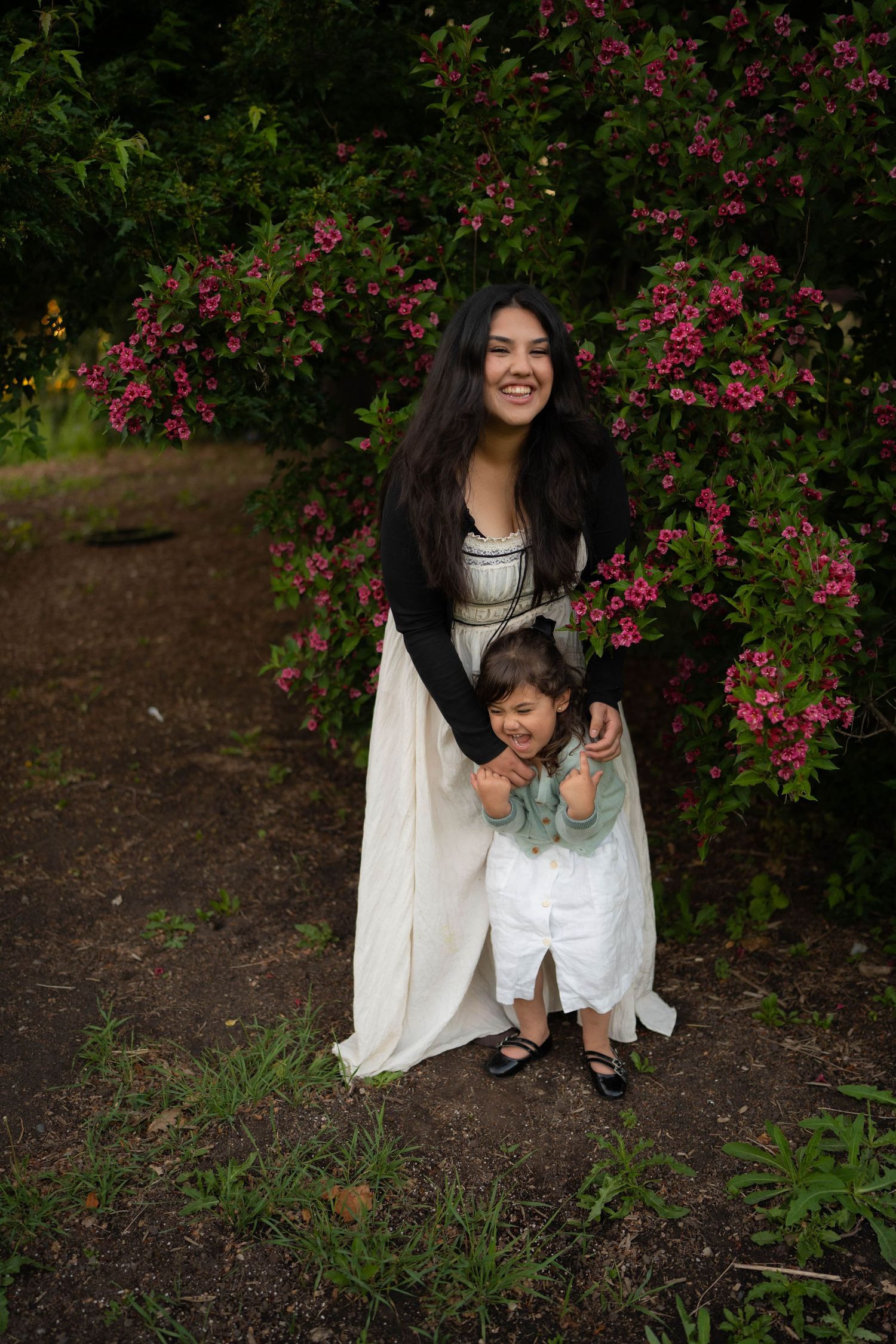 A laughing mother in a cream dress embraces her smiling young daughter in front of a pink flowering bush.