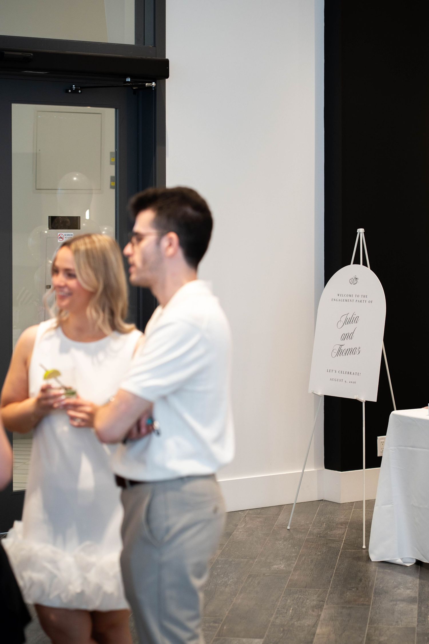A smiling woman in a white dress and a man in a white polo stand near an engagement party welcome sign on an easel.