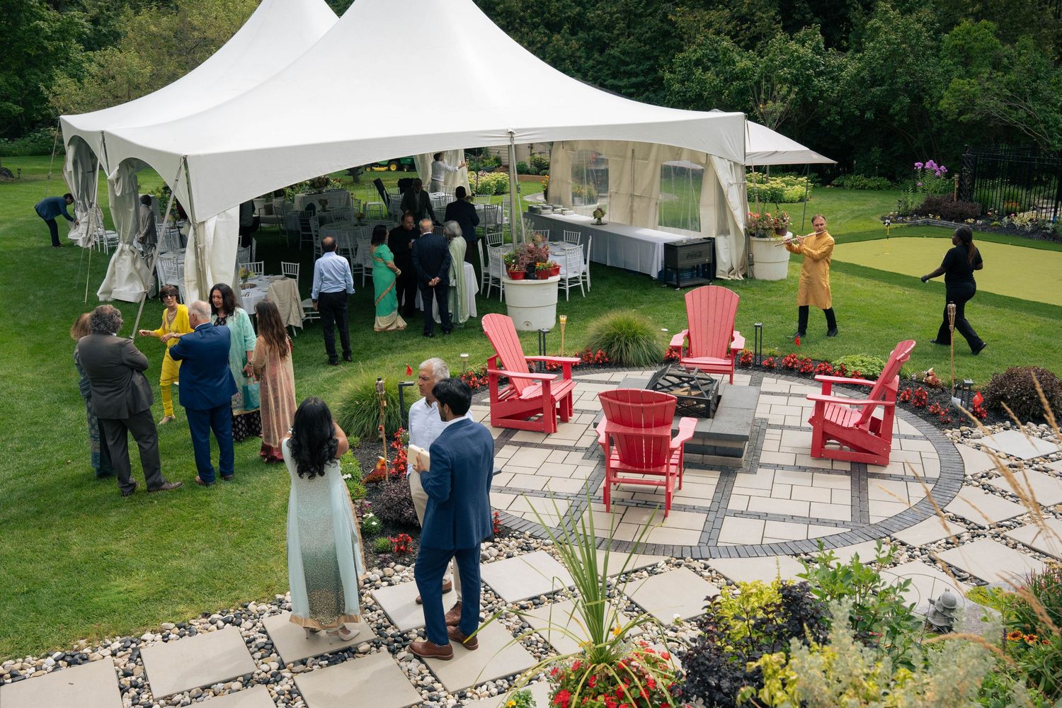 Wedding guests mingle under a white tent and around red Adirondack chairs on a lush lawn with a stone patio.