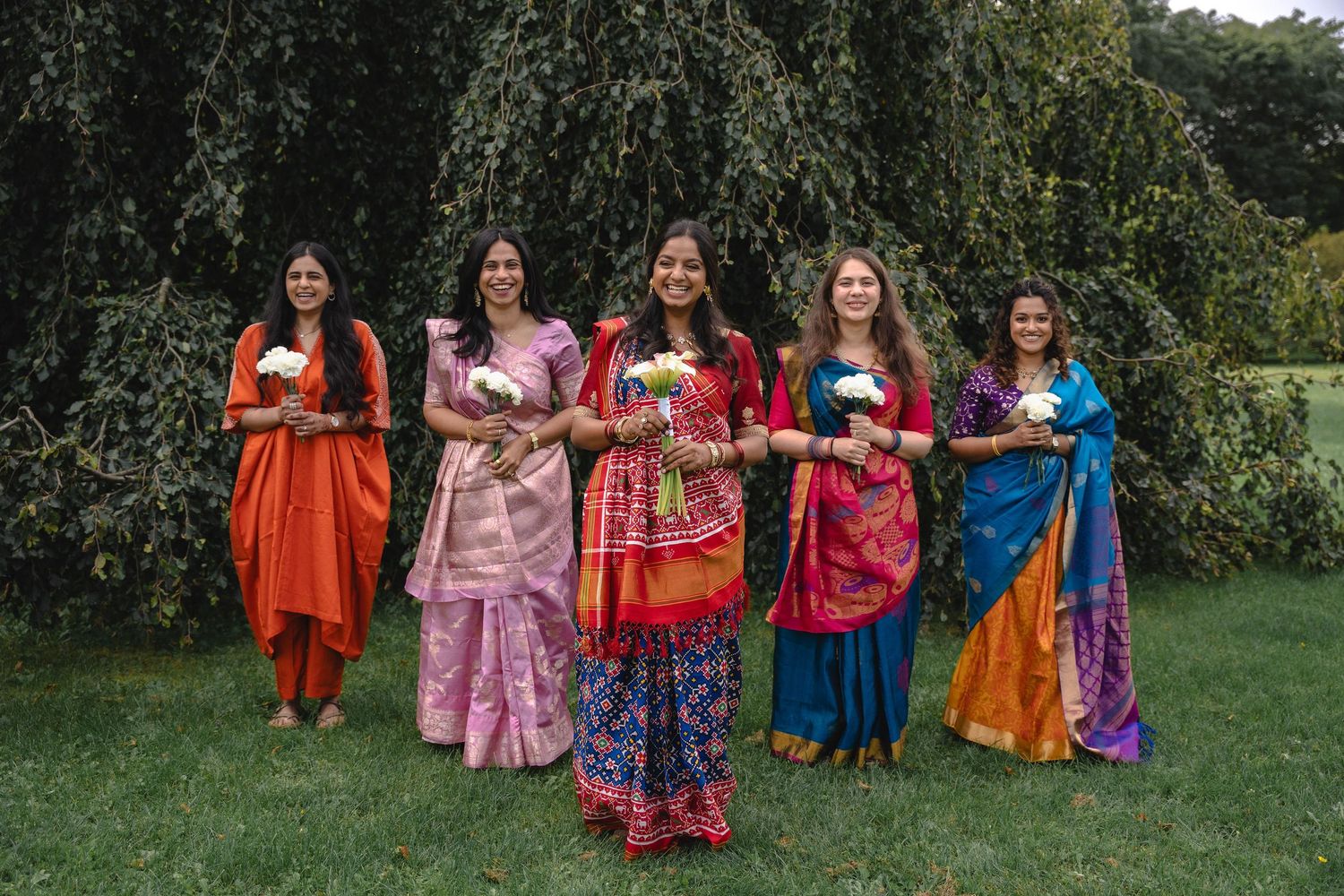 Five women in colorful silk sarees hold white flowers while standing together on a lawn beneath a large weeping tree.