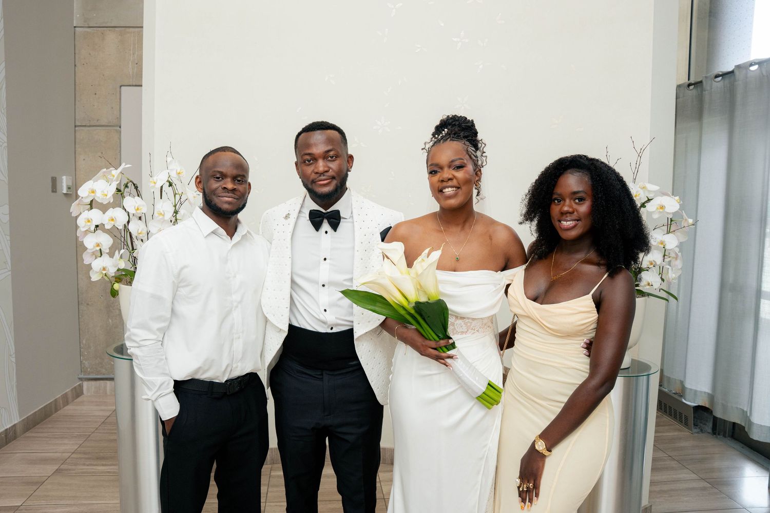 Bride and groom pose with two guests in a bright room, the bride holding a bouquet of white calla lilies.