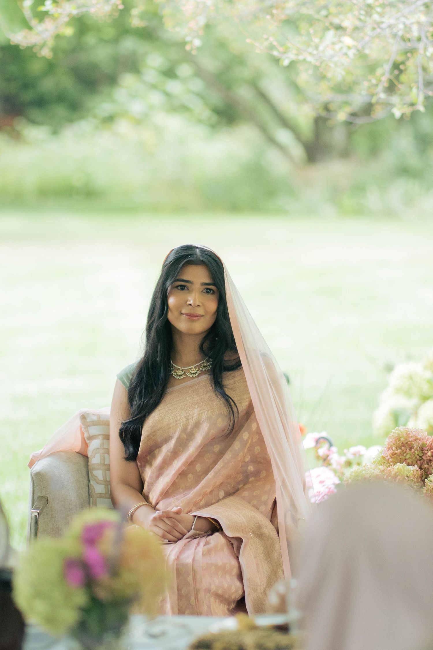 A bride with long dark hair and a peach sari with sheer veil sits smiling on a cushioned seat in a sunlit garden.